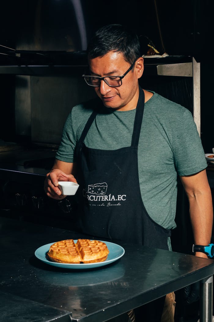 Chef wearing an apron adds final touches to a freshly cooked waffle on a plate in a professional kitchen.